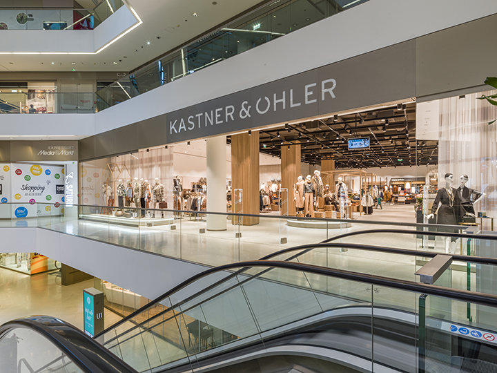 Vista interior de un centro comercial moderno con la tienda Kastner & Öhler. En la fachada de la tienda se ven maniquíes con diferentes atuendos. En primer plano se ven escaleras mecánicas y el centro comercial tiene varios niveles con barandillas de cristal.