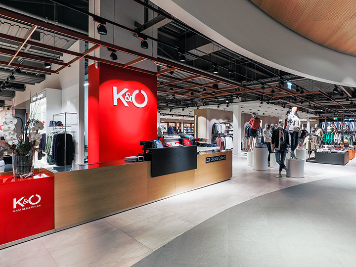 Vista interior de una tienda de ropa moderna con un mostrador rojo brillante que muestra el logotipo de K&Ö. En la tienda se ven varios burros con ropa, con maniquíes y una persona mirando al fondo. El ambiente es espacioso y bien iluminado.
