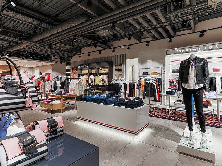 Un interior moderno de una tienda de ropa con diversas prendas. En primer plano, maniquíes exhiben conjuntos de moda. En estanterías se encuentran prendas dobladas y en mesas se exhiben accesorios. Una iluminación brillante realza el diseño moderno de la tienda.