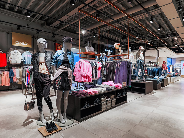 Vista interior de una tienda de ropa con maniquíes vestidos con ropa casual, incluyendo chaquetas y faldas. Varias prendas de vestir, como camisas y jeans, se exhiben en estantes y perchas. La tienda tiene un diseño moderno con iluminación de techo y suelo de baldosas.
