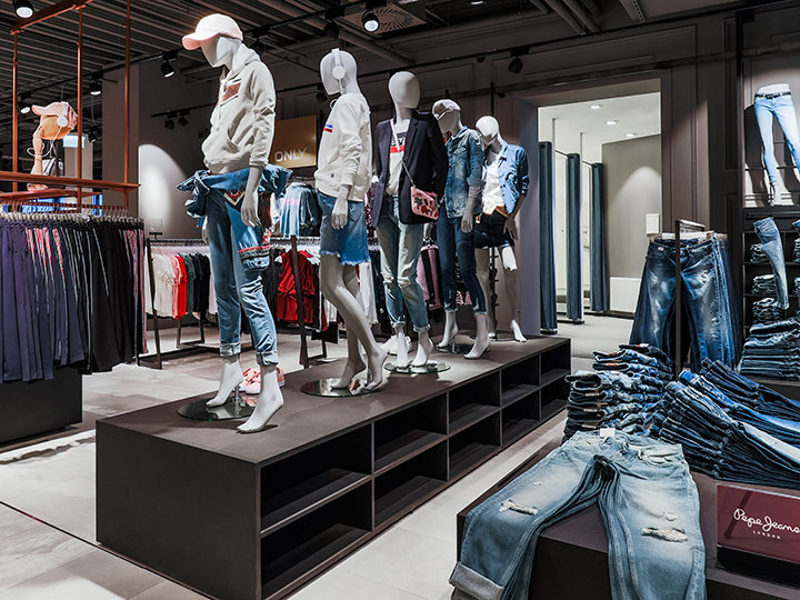 En una tienda de ropa, se exhiben maniquíes con conjuntos informales de mezclilla, que incluyen jeans y chaquetas. Los estantes circundantes contienen varias prendas de vestir, y en un estante se puede ver una pila de jeans doblados. La tienda tiene un diseño moderno.