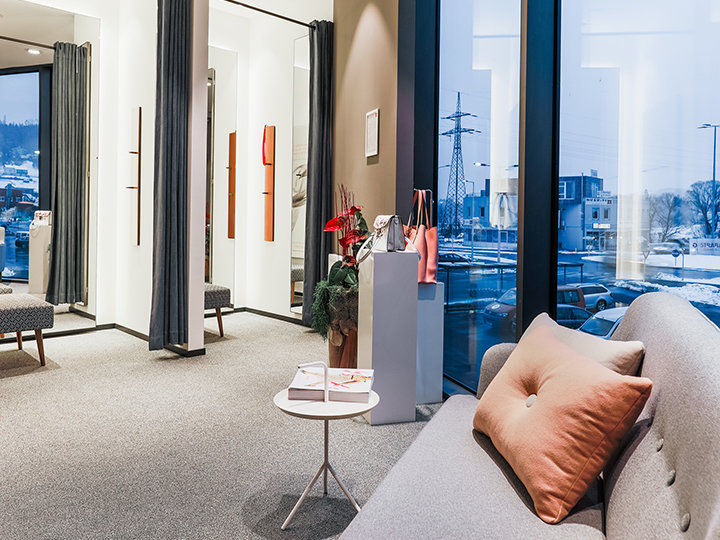 Una moderna y elegante sala de espera con un sofá, una pequeña mesa redonda y grandes ventanas que ofrecen vistas a un paisaje urbano nevado. La habitación está equipada con alfombras, cortinas y plantas decorativas en un pedestal.