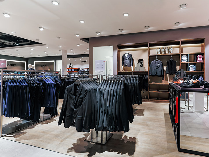 Una tienda de ropa moderna con percheros llenos de trajes colgados ordenadamente. En los estantes del fondo se ven camisas dobladas, blazers y accesorios. La tienda está bien iluminada y tiene un diseño elegante y ordenado.