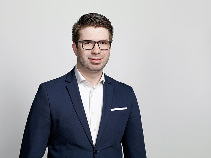Un hombre con gafas, traje azul y camisa blanca está de pie frente a un fondo claro y sencillo. Tiene el pelo corto y castaño y sonríe levemente.