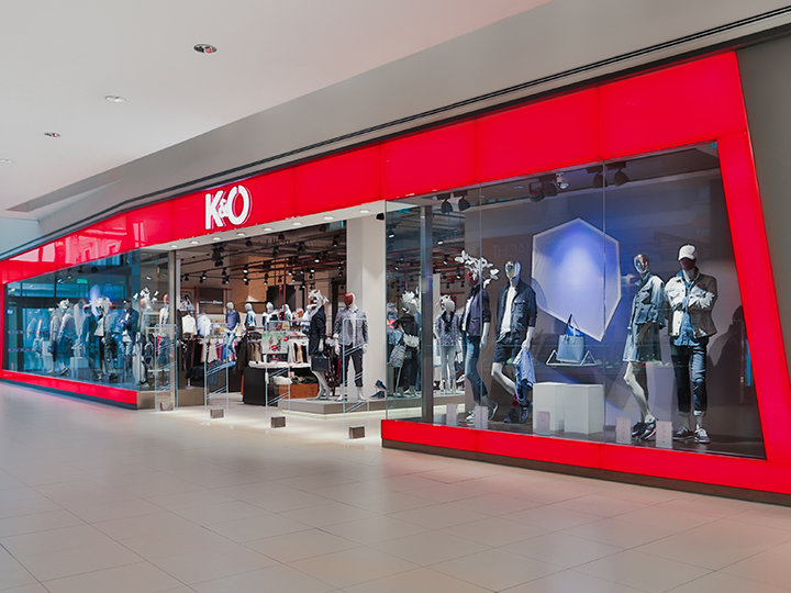 Escaparate con una gran fachada de cristal rojo que exhibe maniquíes vestidos con ropa casual. El interior es claramente visible y muestra percheros. El logo de la tienda muestra una "K&Ö" estilizada.