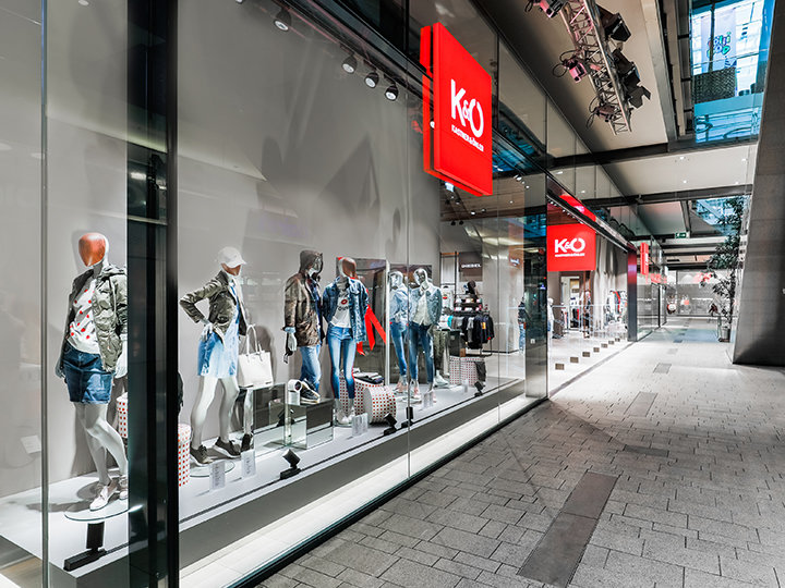 Un escaparate de una tienda de moda que exhibe maniquíes con atuendos casuales de moda, incluyendo chaquetas y pantalones cortos de mezclilla. La tienda tiene grandes ventanales y está ubicada en un moderno centro comercial con arquitectura elegante.