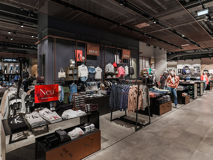 Una tienda de ropa moderna con varias secciones donde se exhiben ordenadamente camisas, chaquetas y accesorios. La tienda se caracteriza por una iluminación brillante, exhibiciones claras y señalización de marca, creando un ambiente de compra elegante y acogedor.