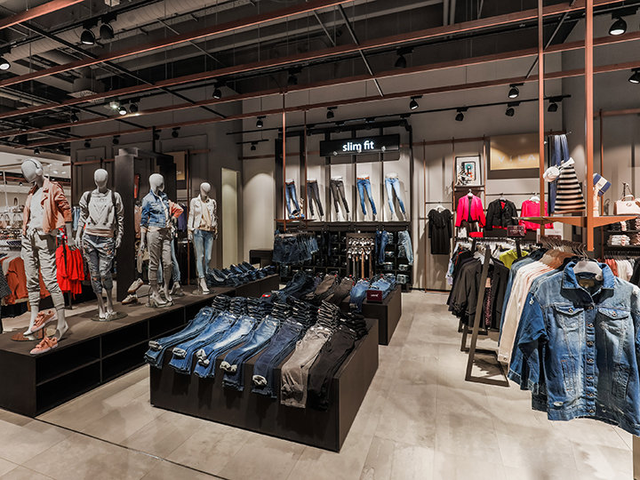 Vista interior de una tienda de ropa con maniquíes que lucen conjuntos de vaqueros. En las mesas de exhibición se presentan vaqueros doblados y en los estantes hay varias prendas de vestir. La tienda tiene un diseño moderno, con iluminación industrial y un letrero que dice "Slim Fit".