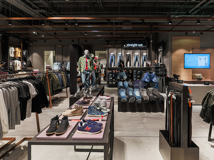 Un interior moderno de una tienda de ropa con varias prendas, incluyendo camisas, chaquetas y vaqueros. Maniquíes exhiben conjuntos, zapatos están dispuestos sobre mesas y se ve una pantalla digital en el fondo. El letrero dice "corte recto".