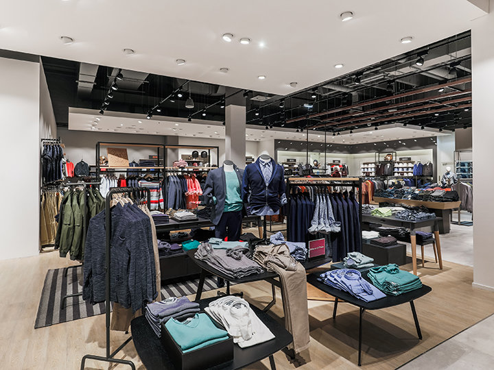 Un interior moderno de una tienda de ropa con estantes y mesas ordenadas que exhiben varias prendas como camisas, chaquetas y pantalones. El espacio está bien iluminado con focos y tiene un diseño limpio y organizado con tonos neutros.