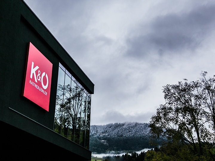 Frente a un cielo nublado, se representa un edificio moderno con un gran letrero rojo que dice "K&O Kastner & Öhler". Colinas cubiertas de nieve y árboles frondosos rodean el edificio, creando un contraste entre elementos urbanos y naturales.