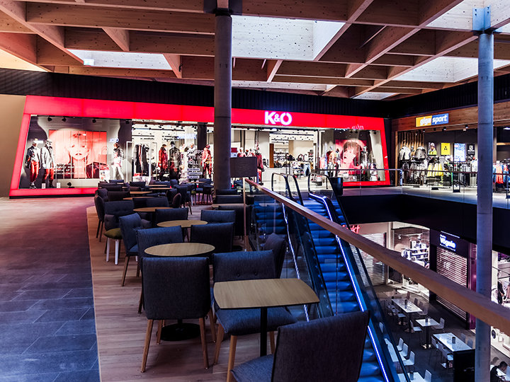 El interior de un moderno centro comercial con una tienda de ropa iluminada en rojo llamada K&O. El espacio incluye áreas de descanso con sillas y mesas, una escalera mecánica y varias otras vitrinas al fondo.