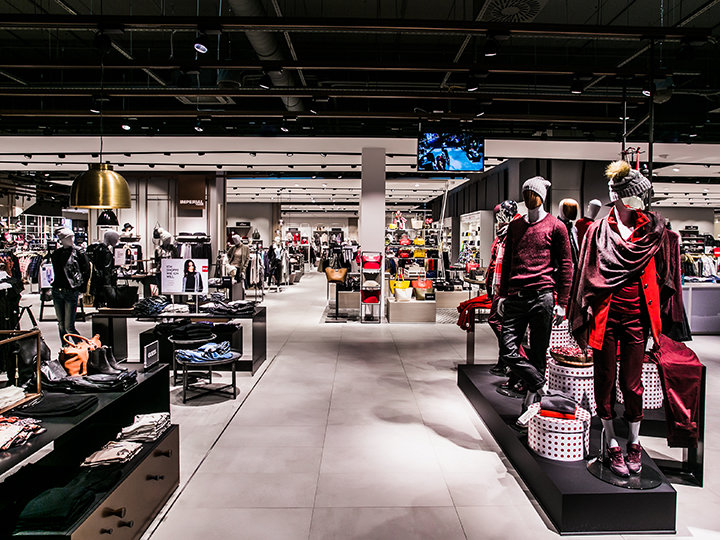 Un interior moderno de una tienda de ropa con maniquíes luciendo atuendos de moda. Estantes y perchas llenos de diversas prendas y accesorios. La tienda está bien iluminada y el espacio es amplio, con la estructura del techo a la vista.