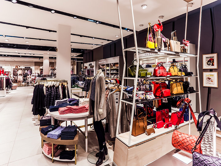 Una tienda de ropa y accesorios con estantes llenos de bolsos coloridos, bufandas y ropa cuidadosamente doblada. Los maniquíes visten atuendos de moda. La tienda está bien iluminada y decorada con un estilo moderno.