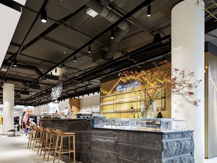 Interior de cafetería moderno con mostrador de piedra, taburetes de madera y plantas ornamentales. El techo es oscuro y cuenta con iluminación sobre rieles, al fondo se ve una zona de estar con gente. Una instalación geométrica dorada en el techo añade un toque elegante al espacio.