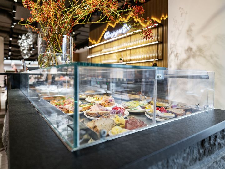 Vitrina de cristal en un restaurante moderno, donde se presentan varios platos de comida, incluyendo carne, queso y verduras. En la parte superior hay ramas decorativas con bayas rojas. Pared de madera con señalización iluminada al fondo.