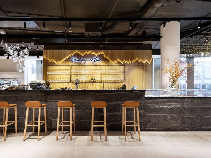 Un bar de champán moderno con una elegante barra de piedra y cinco taburetes marrones. Detrás de la barra, se exhiben copas y botellas en estantes. Un letrero de neón dice "Champagne Bar". Una lámpara decorativa y un arreglo floral añaden elegancia al espacio.