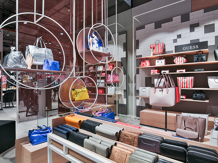 Una elegante exposición de bolsos en venta con una variedad de colores y estilos cuenta con estanterías y perchas redondas. El interior de la tienda, luminoso y moderno, se caracteriza por un llamativo letrero de Guess entre los bolsos ordenados.