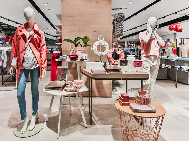 Un escaparate elegante para una tienda de ropa con maniquíes vestidos con ropa casual de primavera. En las mesas se exhiben accesorios como bolsos y joyas. La tienda está bien iluminada y organizada, lo que crea un ambiente de compra moderno y acogedor.
