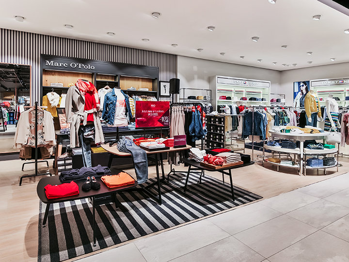 Una tienda de ropa moderna con estantes y exhibidores ordenados con camisas, chaquetas y pantalones coloridos. La tienda está bien iluminada y hay una alfombra de rayas en el suelo. En el fondo se ven carteles de marcas como "Mare O'Polo".