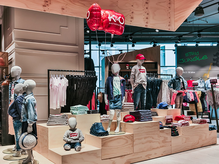 Escaparate de una tienda de ropa infantil con maniquíes vestidos con ropa informal, incluyendo vaqueros y camisas rojas. Los estantes están llenos de ropa doblada y accesorios. Globos rojos cuelgan por encima y en el fondo, un letrero de neón dice "STAY JUNGLE".