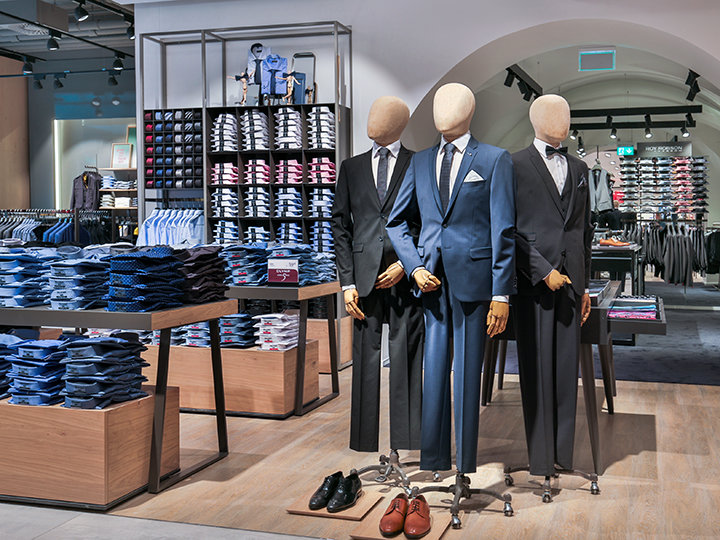 Un escaparate en una tienda de ropa con tres maniquíes vestidos con trajes formales. En el entorno se ven camisas y accesorios apilados ordenadamente. La tienda tiene un interior moderno con iluminación suave y mercancía bien organizada.