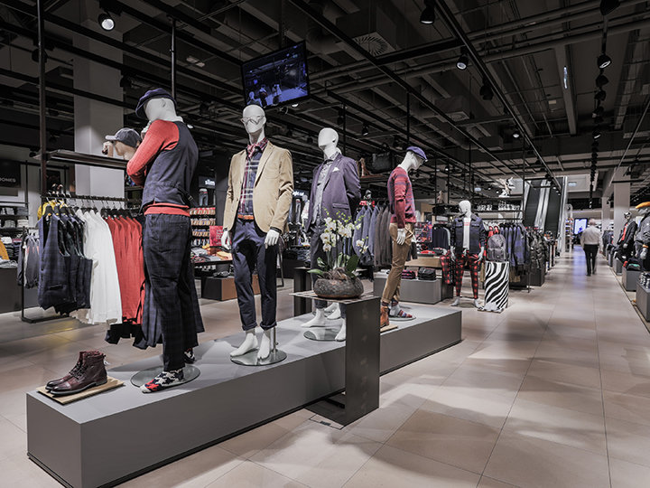 Un escaparate en una tienda de ropa con maniquíes vestidos con atuendos casuales y de noche. La tienda es espaciosa, con percheros de ropa y accesorios bajo una iluminación brillante al fondo. Una persona camina por el pasillo.