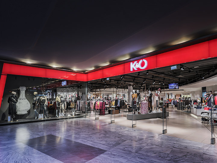Escaparate de una tienda de moda K&O con maniquíes que exhiben diversos conjuntos tras grandes ventanales. La tienda tiene un diseño elegante y moderno, y un letrero rojo brillante con el logotipo de K&O colocado de forma destacada sobre la entrada.