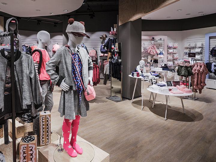 Una tienda de ropa infantil con maniquíes luciendo atuendos coloridos, incluyendo un sombrero y leggings rosas. Varias prendas de vestir y accesorios se exhiben en estantes y perchas. El suelo es de madera y la tienda tiene una decoración interior moderna.