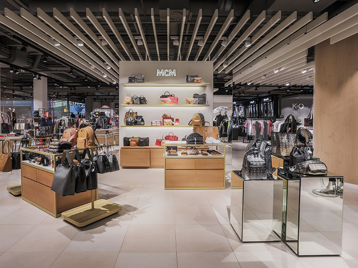 Un interior de tienda moderno donde se exhibe una variedad de bolsos de lujo en expositores de madera y vidrio. En los estantes de la pared se pueden ver más bolsos. El techo tiene un diseño geométrico y la iluminación es brillante, resaltando la mercancía.