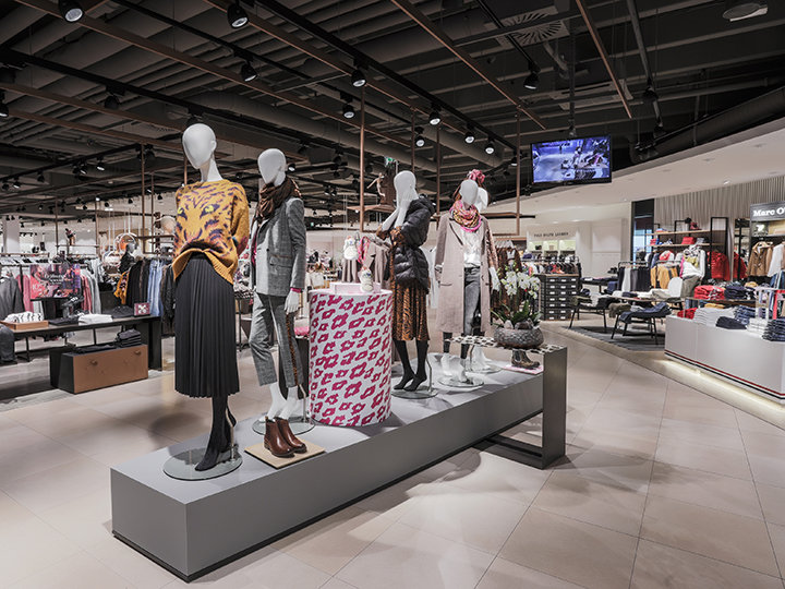 Un interior elegante de una tienda de ropa con maniquíes luciendo conjuntos de moda. La tienda tiene una decoración moderna, percheros y una gran caja estampada. La iluminación del techo resalta varias exhibiciones y se ve una pantalla al fondo.