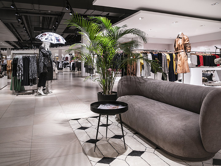 Un interior moderno de una tienda de ropa con percheros, incluyendo abrigos y chaquetas. En primer plano hay un sofá gris y una mesa redonda negra junto a una planta en maceta. En el fondo, un maniquí sostiene un paraguas.