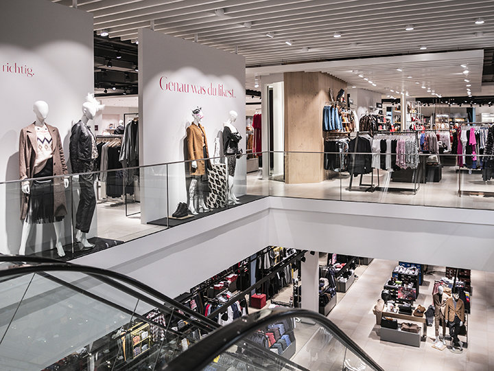 Un interior moderno de una tienda de ropa con maniquíes vestidos con ropa casual exhibidos en una plataforma. Percheros y estanterías están ordenados en dos pisos, visibles desde la escalera mecánica en primer plano.