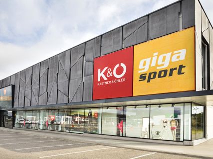 Außenansicht eines modernen Gebäudes mit großen Schildern für „Kastner & Öhler“ in Rot und „Giga Sport“ in Gelb. Im Erdgeschoss sind große Schaufenster mit Sport- und Modeartikeln zu sehen. Der Himmel ist bedeckt.