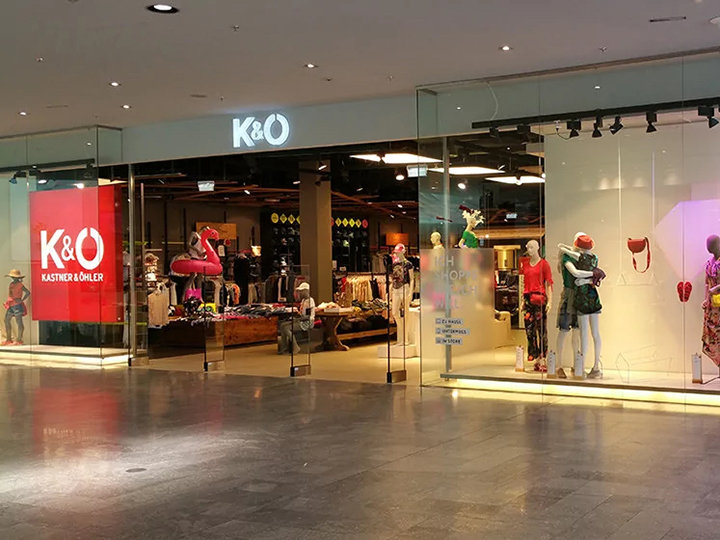 Escaparate de Kastner & Öhler con grandes ventanales que muestran maniquíes vestidos. En el interior de la tienda se ven percheros y una selección de ropa. El logotipo de K&O brilla claramente visible sobre la entrada.