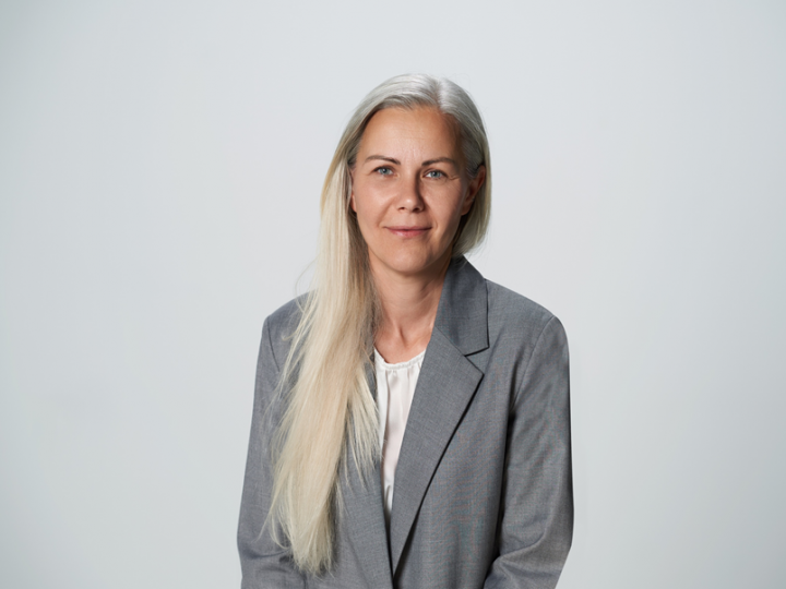 Una persona con cabello largo y rubio, un blazer gris claro y una blusa blanca está de pie frente a un fondo sencillo de color gris claro. Tiene una expresión facial neutra y mira a la cámara.