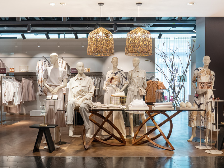 En una tienda de ropa, varios maniquíes lucen atuendos neutros y modernos. Están rodeados de iluminación suave, decoración moderna y prendas de vestir ordenadas en perchas y mesas. Sobre ellos cuelgan lámparas colgantes tejidas.