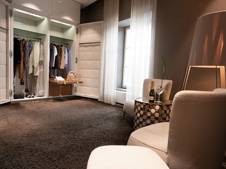 Una habitación moderna y elegante con un armario abierto que exhibe ropa y un bolso. Dos cómodas sillas están frente a una mesa redonda con una lámpara decorativa. Una botella de champán y copas están sobre la mesa y la luz entra a través de las cortinas transparentes.
