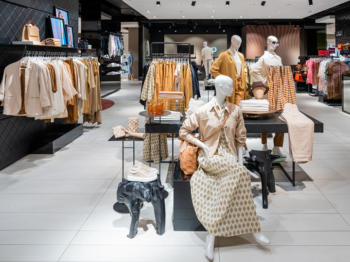 Interior de una tienda de ropa con prendas en tonos neutros en perchas y maniquíes. Se exhiben camisas, chaquetas, vestidos y accesorios como bolsos y sombreros. Iluminación brillante, con un diseño moderno y ordenado.