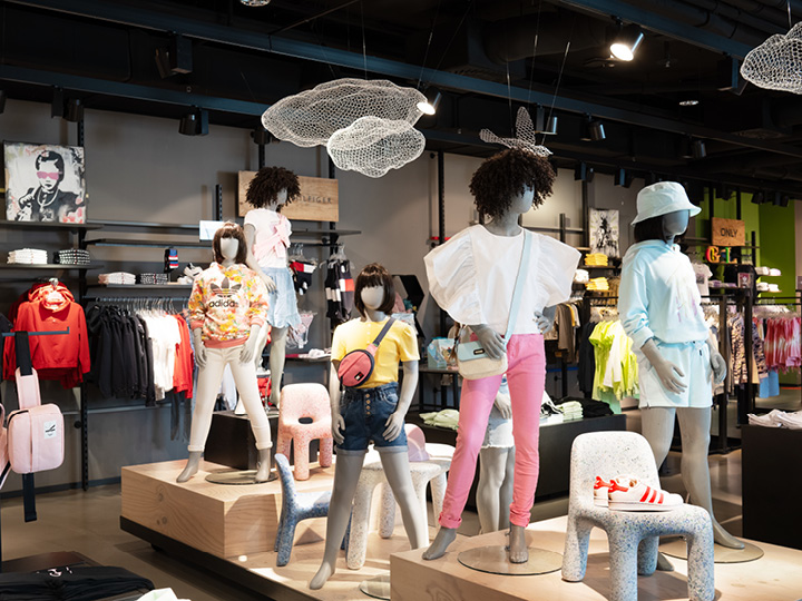 En una tienda de ropa de moda, se exhiben maniquíes con atuendos coloridos e informales. La tienda ofrece una variedad de prendas de vestir y accesorios sobre un fondo elegante y moderno con elementos decorativos artísticos que cuelgan del techo.