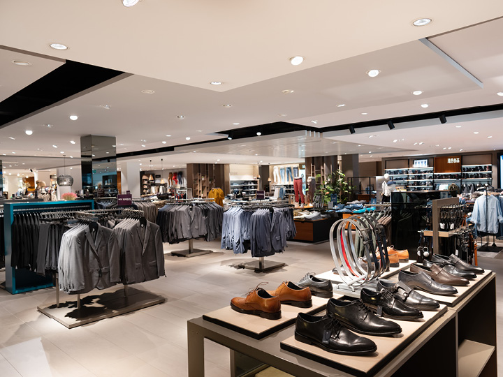 Un interior espacioso de una tienda de ropa con trajes ordenados en perchas, zapatos elegantes en una mesa de exhibición y accesorios como cinturones. La tienda está bien iluminada y decorada de forma moderna, lo que crea un ambiente de compras exclusivo.