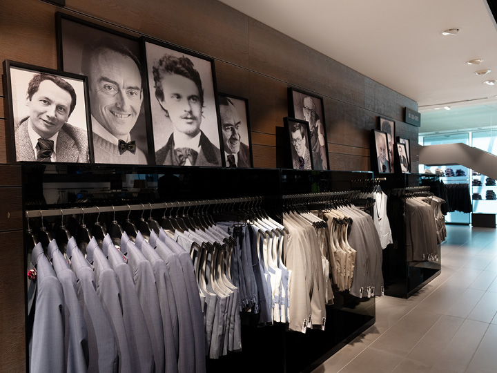 En una tienda de ropa, cuelgan trajes ordenadamente en varios tonos de azul y beige. Sobre los trajes, cuelgan retratos en blanco y negro de varios hombres enmarcados en una pared de madera.