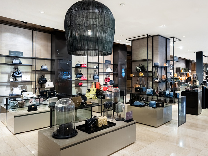 Una tienda minorista de lujo exhibe bolsos de diseño en elegantes estantes y mostradores. El espacio cuenta con iluminación moderna, tonos neutros y una gran lámpara de techo negra, creando un ambiente de compras elegante.