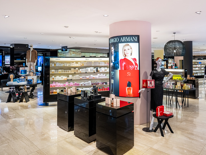 Un lujoso centro comercial presenta varias marcas de perfumes, incluyendo Giorgio Armani. La luz brillante resalta modernos expositores negros llenos de productos. En el fondo, estantes llenos de más artículos de belleza.