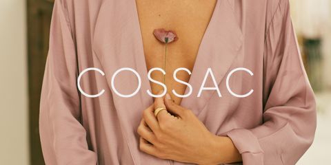 Una persona con una bata de seda de color rosa pálido sostiene un tallo de flor rosa entre sus dedos cerca de su pecho. La palabra "COSSAC" está superpuesta en la imagen en letras blancas.