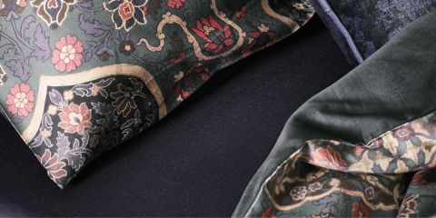 Primer plano de una cama con almohadas oscuras con estampado floral y una manta estampada a juego. La tela presenta intrincados diseños en tonos rojos, rosas y verdes que contrastan con un fondo oscuro. La tela parece lujosa y suave.