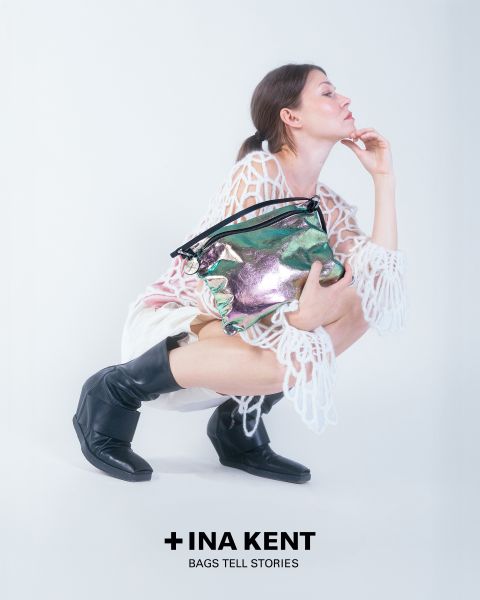 Sobre un fondo blanco, una persona está sentada en cuclillas, vestida con un conjunto de encaje blanco y botas negras. Sostiene un bolso brillante y colorido en la mano. Abajo se puede ver el texto "+ INA KENT BOLSOS CUENTAN HISTORIAS".