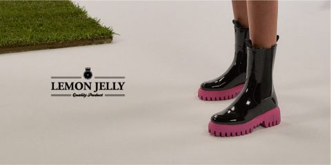 Persona con brillantes botas negras con suelas rosas sobre suelo blanco, junto a una mancha de hierba verde. A la izquierda está el texto "Producto de calidad Lemon Jelly".