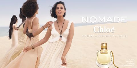 Cuatro mujeres caminan con vestidos ondeantes a través de un paisaje desértico. A la derecha se ve el texto "Nomade Chloé" y un frasco de perfume que promociona la fragancia. La escena transmite una sensación de libertad y elegancia natural.