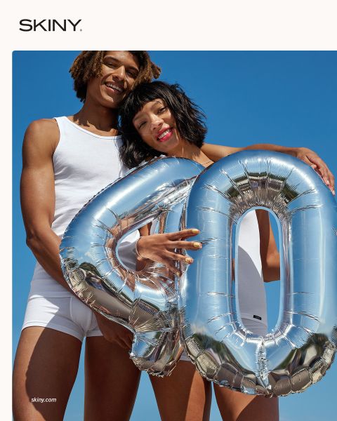 Dos personas en ropa interior blanca sostienen sonriendo un globo plateado con el número "40" frente a un cielo azul brillante.